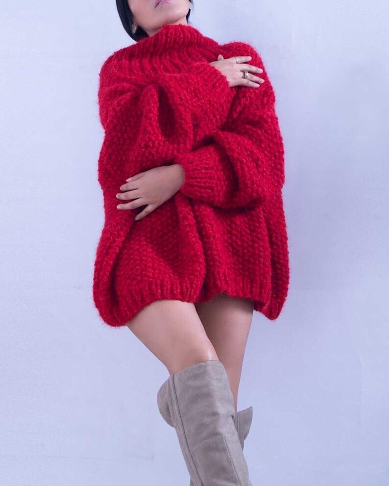 May include: A bright red, chunky knit sweater featuring a high turtleneck collar, long sleeves with ribbed cuffs, and a loose, oversized fit. The garment is crafted from a thick, textured yarn with a visible weave, providing a voluminous silhouette.