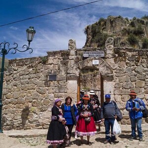 May include: A group of people stand in front of a stone archway with a sign that reads "Way to the Sanctuary of the Holy Cross". The archway is made of rough-hewn stone and is surrounded by a stone wall. The people are wearing traditional clothing and hats.