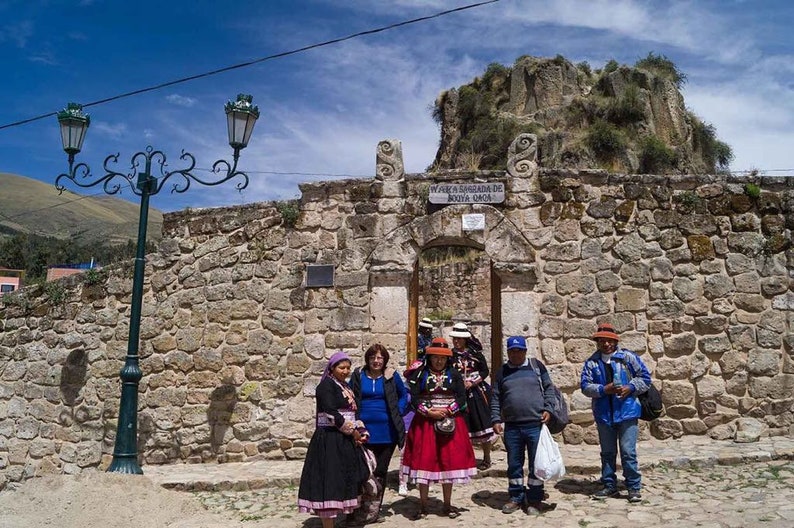 May include: A group of people stand in front of a stone archway with a sign that reads "Way to the Sanctuary of the Holy Cross". The archway is made of rough-hewn stone and is surrounded by a stone wall. The people are wearing traditional clothing and hats.