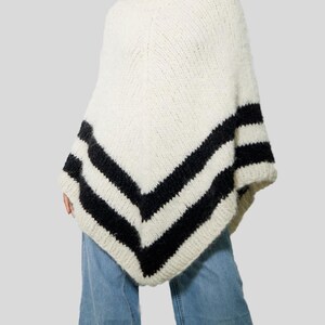 Pu&ograve; includere: Un poncho in maglia grossa color bianco sporco con un orlo a forma di V. Il capo &egrave; decorato con due spesse strisce orizzontali nere vicino al bordo inferiore, creando un netto contrasto con il filato testurizzato color crema.