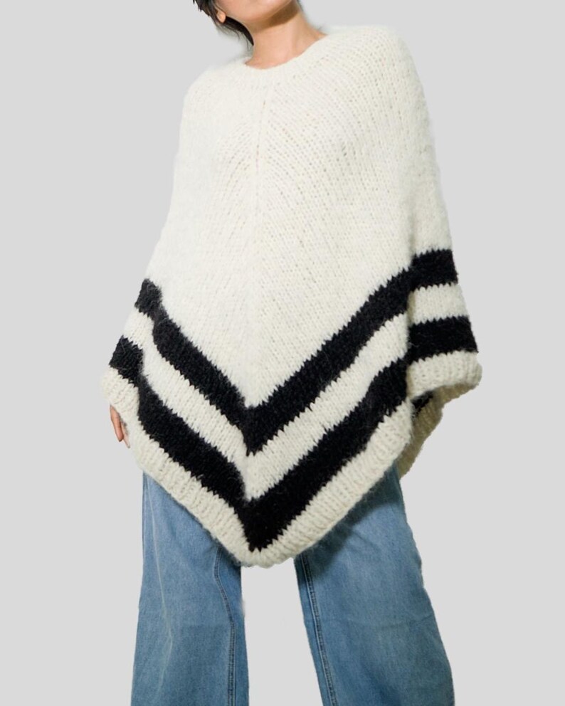 Pu&ograve; includere: Un poncho in maglia grossa color bianco sporco con un orlo a forma di V. Il capo &egrave; decorato con due spesse strisce orizzontali nere vicino al bordo inferiore, creando un netto contrasto con il filato testurizzato color crema.