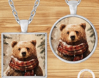 May include: Two silver-toned pendant necklaces. One pendant is square, the other is round. Both feature a brown bear wearing a red and black plaid scarf. The bear is set against a snowy forest background.