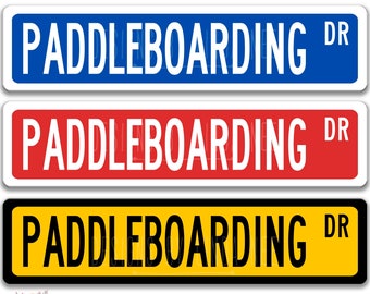 以下が含まれることがあります： 青、赤、黄色の背景に白い文字で「PADDLEBOARDING DR」と書かれたアルミ製の標識が3つあります。標識は4インチ×18インチで、アメリカ製です。