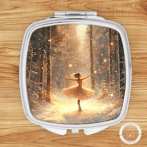 Może przedstawiać: Srebrne lusterko kompaktowe z zimową sceną leśną i baletnicą w białej sukience. Grafika przedstawia zaśnieżony las z baletnicą tańczącą pośrodku, z ciepłą, złotą poświatą. Lusterko ma kwadratowy kształt z zaokrąglonymi krawędziami.
