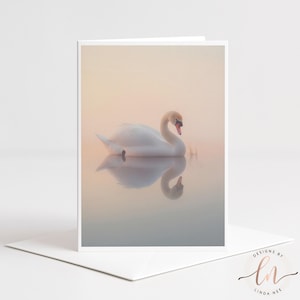 Könnte beinhalten: Eine Grußkarte mit einem ruhigen Bild eines weißen Schwans, der anmutig auf ruhigem Wasser schwimmt. Die Spiegelung des Schwans spiegelt sich im Wasser wider, vor einem Hintergrund in sanften Pastellfarben. Die Karte befindet sich auf einem weißen Umschlag.