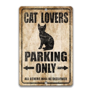 Pu&ograve; includere: Un cartello in metallo in stile vintage con uno sfondo marrone invecchiato. Il cartello recita "Cat Lovers Parking Only" con una silhouette nera di un gatto seduto al centro. Il testo "All others will be declawed" &egrave; stampato sotto il gatto.