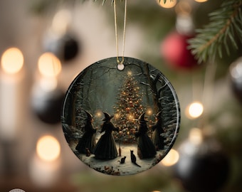 May include: Round Christmas ornament featuring a winter forest scene with four witches gathered around a decorated Christmas tree. The witches wear black robes and hats, holding wands. Two black cats are in the foreground. The ornament hangs from a gold string.