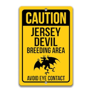 May include: Yellow caution sign with black text and a black silhouette of a bat-like creature. The sign reads "Caution Jersey Devil Breeding Area Avoid Eye Contact."
