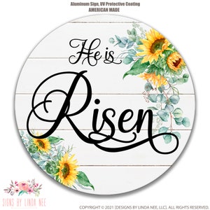 May include: A white wooden round sign with a whitewashed woodgrain background. The sign features a black cursive font that reads "He is Risen". The sign is decorated with a floral arrangement of sunflowers and eucalyptus leaves.