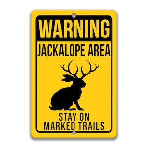 May include: Yellow warning sign with black text and a silhouette of a jackalope. The sign reads "WARNING JACKALOPE AREA STAY ON MARKED TRAILS".