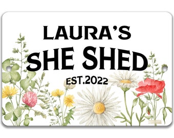 Könnte beinhalten: Ein weißes Schild mit schwarzem Text, der "Laura's She Shed Est. 2022" lautet. Das Schild ist mit Aquarellblumen in den Farben Rosa, Gelb und Rot verziert.