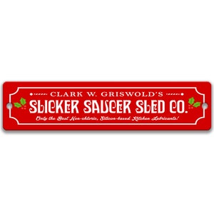 Puede incluir: Un letrero rojo con texto blanco que dice "... CLARK W. GRISWOLD'S ... SLICKER SAUCER SLED CO. Only the Best Non-chloric, Silicon-based Kitchen Lubricants!" con una rama de acebo verde a cada lado.
