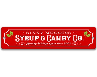 May include: Red rectangular sign with white text that reads "Ninny Muggins Syrup & Candy Co. Keeping holidays hyper since 2003."  The sign has a green holly leaf design on each side.