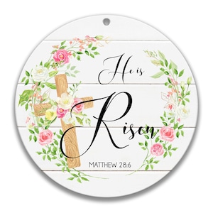 May include: A white wooden circle with a floral wreath and a wooden cross. The text "He is Risen" is written in black cursive script with the scripture reference "Matthew 28:6" below.
