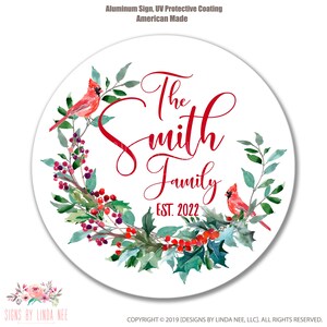 Pu&ograve; includere: Un cartello circolare di metallo bianco con una ghirlanda ad acquerello rossa con agrifoglio, bacche e due cardinali rossi. Il testo "The Smith Family EST. 2022" &egrave; scritto in lettere corsive rosse.