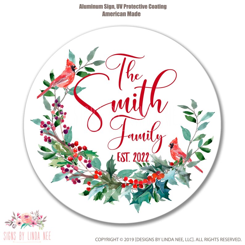 Pu&ograve; includere: Un cartello circolare di metallo bianco con una ghirlanda ad acquerello rossa con agrifoglio, bacche e due cardinali rossi. Il testo "The Smith Family EST. 2022" &egrave; scritto in lettere corsive rosse.