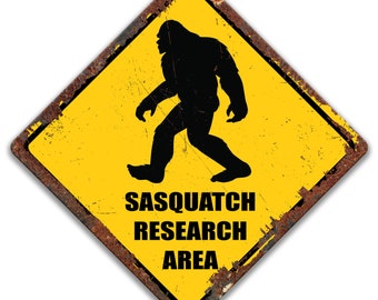 May include: A yellow diamond-shaped metal sign with a black silhouette of a Sasquatch walking. The sign has the words "SASQUATCH RESEARCH AREA" in black text. The sign has a rusty brown border and a distressed appearance.