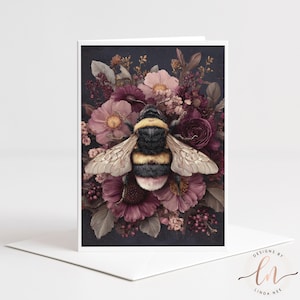 Pode incluir: Cartão de felicitações com uma ilustração detalhada de um abelha com listras pretas e amarelas, em um fundo de flores bordô e rosa. O cartão tem uma borda branca e é colocado em um envelope branco.