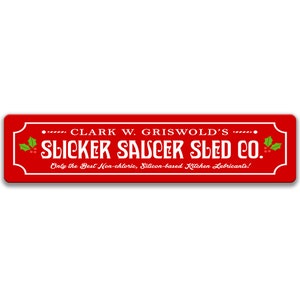 Puede incluir: Un letrero rojo con texto blanco que dice "Clark W. Griswold's Slicker Saucer Sled Co. Only the Best Non-chloric, Silicon-based Kitchen Lubricants!" El letrero tiene una rama de acebo con bayas rojas a cada lado.