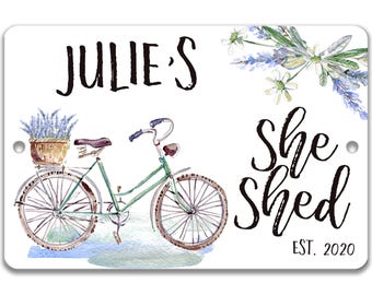 Op de afbeelding: Een rechthoekig metalen bord met een aquarelillustratie van een groene fiets met een mand vol lavendel. De tekst luidt JULIE'S she shed EST. 2020 in zwarte letters. Het ontwerp bevat florale accenten in de rechterbovenhoek.