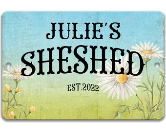 May include: A blue and green sign with white daisies and the text "Julie's Sheshed Est. 2022".