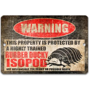 May include: A rectangular metal sign with a distressed, vintage look. The sign features a red "WARNING" oval at the top. Below, text reads "THIS PROPERTY IS PROTECTED BY A HIGHLY TRAINED RUBBER DUCKY ISOPOD." A cartoon isopod is on the right.