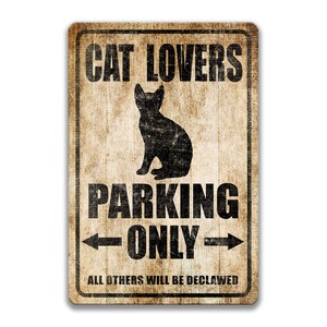 Pu&ograve; includere: Un cartello in metallo in stile vintage con uno sfondo marrone usurato e testo nero. Il cartello recita "Cat Lovers Parking Only" con una silhouette di un gatto seduto sotto il testo. Le frecce puntano a sinistra e a destra del testo "Only". La parte inferiore del cartello recita "All others will be declawed".