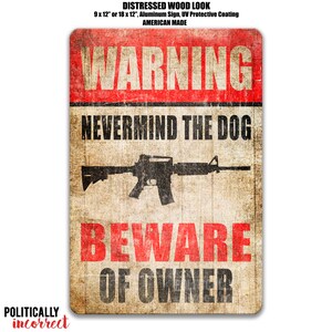 May include: A rectangular aluminum sign with a distressed wood look. The sign features the text "WARNING" in red, "NEVERMIND THE DOG" and "BEWARE OF OWNER" in black. A black rifle graphic is in the center. The sign is 9 x 12 inches or 18 x 12 inches.