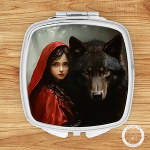 May include: A silver compact mirror featuring a digital illustration of a young woman in a red hooded cloak and a black wolf. The artwork has a fairytale theme, with the woman looking directly at the viewer. The compact is square with rounded corners.