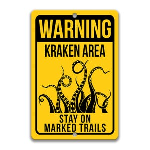 Puede incluir: Señal de advertencia amarilla con texto negro que dice "WARNING KRAKEN AREA STAY ON MARKED TRAILS". La señal presenta una silueta negra de un pulpo con tentáculos.