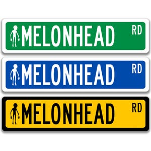 May include: Three street signs reading "MELONHEAD RD". The top sign is green, the middle sign is blue, and the bottom sign is yellow. Each sign has a white border and a small alien-like figure.
