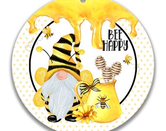 Op de afbeelding: Een ronde, witte ornament met een gele en zwarte bij-gnome die een honingpot met een bij erop vasthoudt. De achtergrond is wit met gele stippen en de tekst "Bee Happy" is in zwart geschreven.