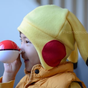 May include: A child wearing a yellow Pikachu hat with red ear accents and a black tail holds a red and white Pokeball. The hat has the text "Pikamomodesign" on the side.