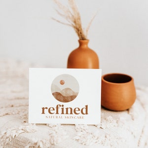May include: A white card with a logo for a natural skincare company called "refined". The logo features a stylized image of a mountain range with a sun in the sky. The card is sitting on a white, fluffy surface.