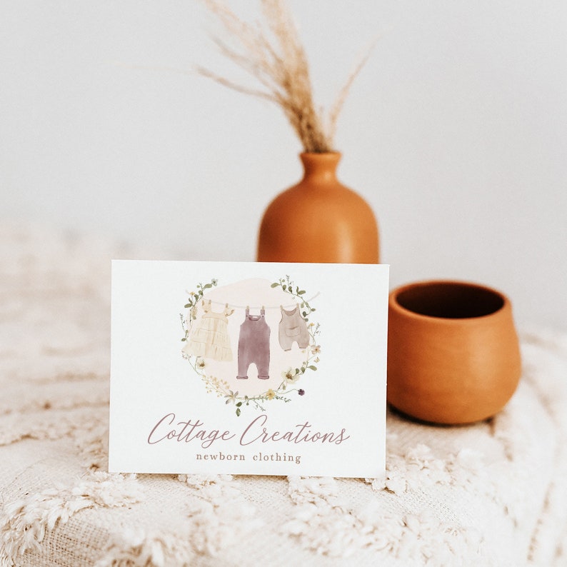 May include: A white card with a logo for a business called "Cottage Creations" that specialises in newborn clothing. The logo features a watercolour illustration of three baby clothes hanging on a clothesline with a floral wreath around them.