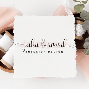 May include: A white card with the text "julia bernard INTERIOR DESIGN" in black cursive script. The card is on a white background with pink fabric and a wooden bowl with white spools of thread.