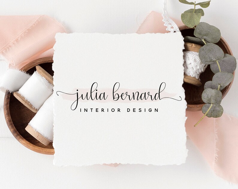 May include: A white card with the text "julia bernard INTERIOR DESIGN" in black cursive script. The card is on a white background with pink fabric and a wooden bowl with white spools of thread.