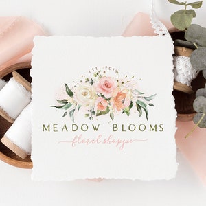 以下が含まれることがあります： ピンクと白のバラ、緑、そして「Est. 2014」の文字が特徴の「Meadow Blooms floral shoppe」の水彩フローラルロゴデザイン。 ロゴは、テクスチャ加工された白い紙の上にあり、背景にはリボンのスプールとユーカリの葉があります。