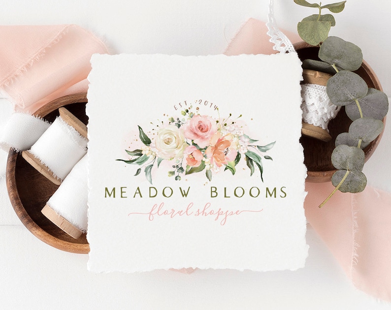 以下が含まれることがあります： ピンクと白のバラ、緑、そして「Est. 2014」の文字が特徴の「Meadow Blooms floral shoppe」の水彩フローラルロゴデザイン。 ロゴは、テクスチャ加工された白い紙の上にあり、背景にはリボンのスプールとユーカリの葉があります。