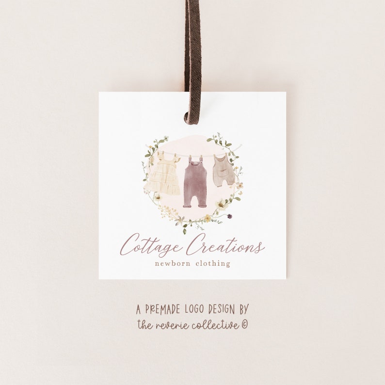 May include: A premade logo design for a business called "Cottage Creations" that sells newborn clothing. The logo features a watercolour illustration of three baby clothes hanging on a clothesline with a floral wreath around them. The text "Cottage Creations" is written in cursive below the illustration, and the text "newborn clothing" is written in a smaller font below that.