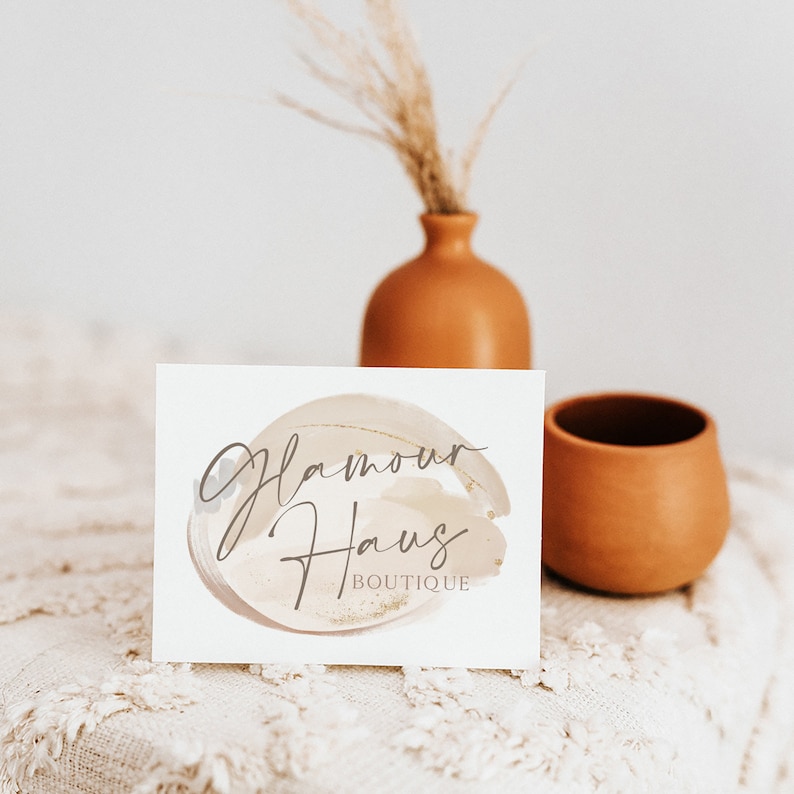May include: A white card with the text "Glamour Haus Boutique" in a gold and gray script font on a beige watercolor background. The card is sitting on a white crocheted surface with a terracotta vase and a terracotta cup in the background.
