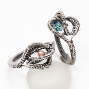 May include: Two silver wire rings with a pink pearl and a blue iridescent gemstone. The rings are intertwined and are on a white surface.