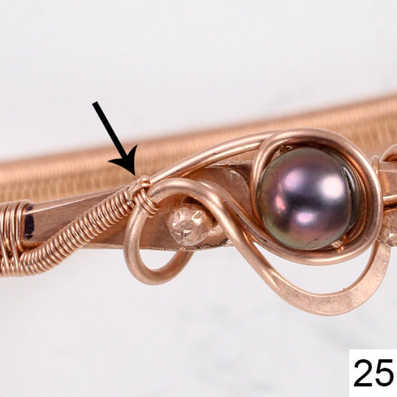 K&ouml;nnte beinhalten: Nahaufnahme eines mit Kupferdraht umwickelten Armbands mit einer dunkelgrauen Perle. Der Draht ist um die Perle gewickelt und wird dann weiter um das Armband gewickelt. Der Draht ist an der Stelle, an der er um die Perle gewickelt ist, verdreht.