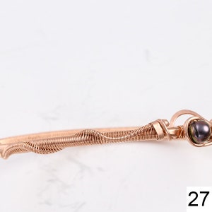 K&ouml;nnte beinhalten: Ein ros&eacute;goldenes, drahtgewickeltes Armband mit einer dunkelgr&uuml;nen Perle. Das Armband besteht aus d&uuml;nnem Draht, der verdreht und miteinander verflochten ist, um ein einzigartiges und filigranes Design zu schaffen.