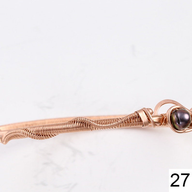 K&ouml;nnte beinhalten: Ein ros&eacute;goldenes, drahtgewickeltes Armband mit einer dunkelgr&uuml;nen Perle. Das Armband besteht aus d&uuml;nnem Draht, der verdreht und miteinander verflochten ist, um ein einzigartiges und filigranes Design zu schaffen.