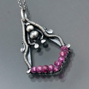 May include: A silver pendant necklace with a delicate, intricate design featuring a cluster of small silver beads and a row of pink gemstones.