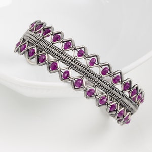 Puede incluir: Un brazalete de alambre plateado envuelto con un diseño geométrico y acentos de piedras preciosas púrpuras.