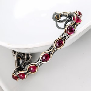 May include: A silver and gold wire wrapped bracelet with red gemstones. The bracelet is made with a delicate wire design and features a series of round, red gemstones.