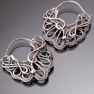 May include: A pair of silver hoop earrings with intricate wirework designs. The earrings are made of silver wire that is twisted and woven together to create a delicate and intricate pattern.
