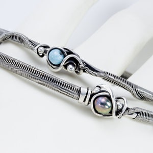 K&ouml;nnte beinhalten: Zwei silberne, drahtgewickelte Armb&auml;nder mit einem blauen Edelstein und einer schwarzen Perle. Die Armb&auml;nder liegen auf einer wei&szlig;en Oberfl&auml;che.